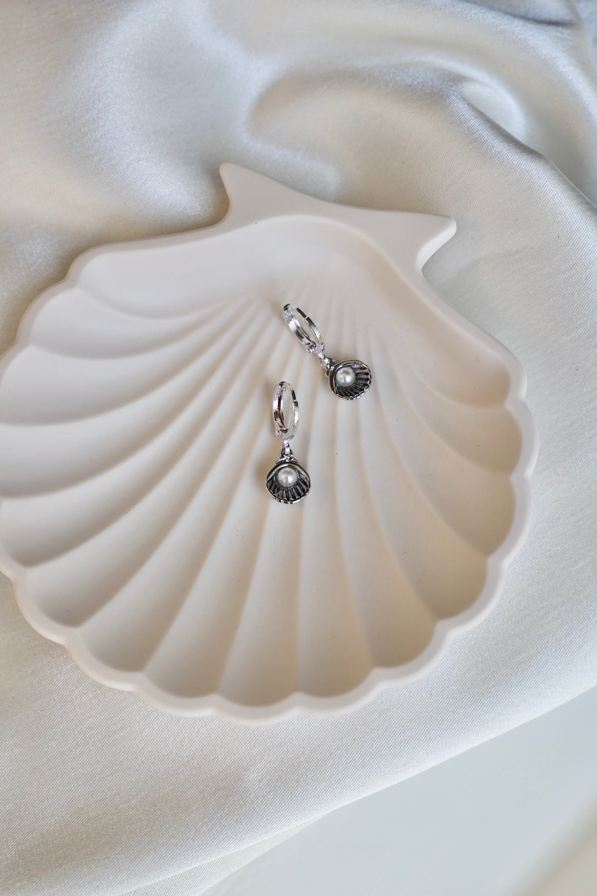 Two silver shell-shaped earrings with pearls are resting on a white, shell-shaped dish. The dish is placed on a soft, light-colored fabric background.