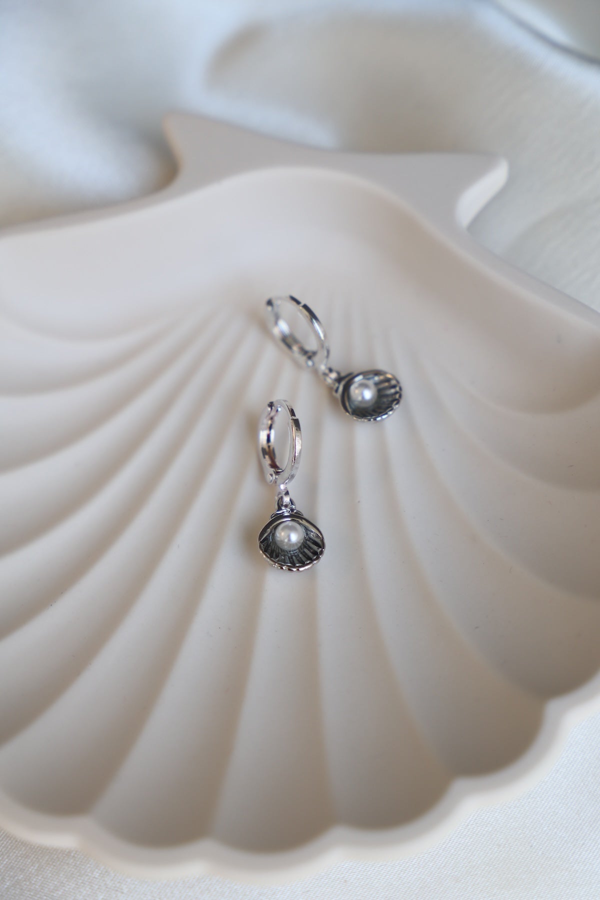 A pair of silver earrings shaped like shells, each containing a pearl, rests on a white, shell-shaped dish. The background is soft and light-colored, creating a serene atmosphere.