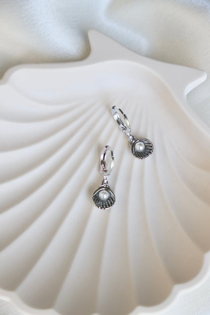 Two silver earrings shaped like shells, featuring small pearls, rest inside a white shell-shaped dish. The background includes a soft, lightly textured fabric.