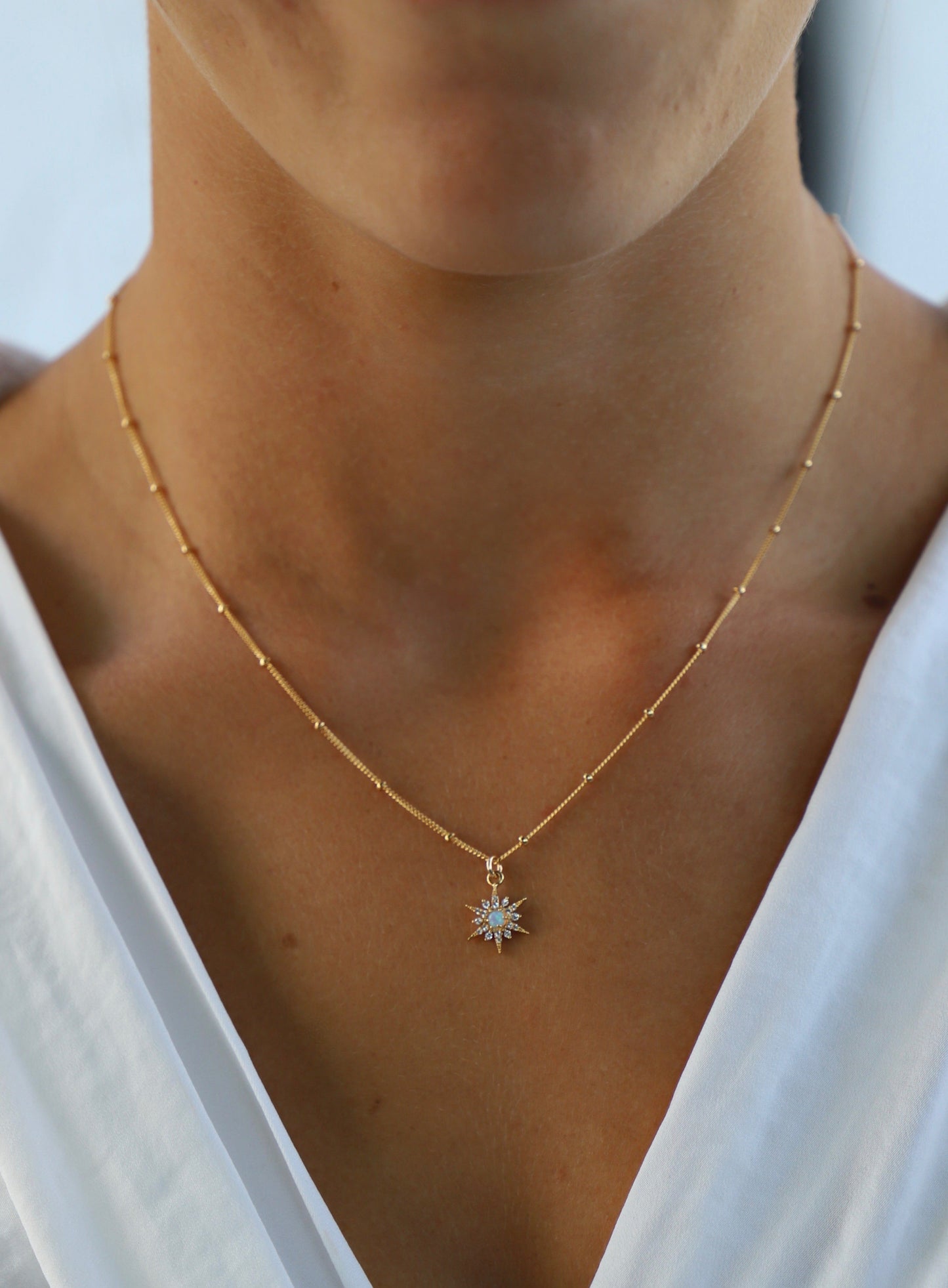 A gold chain necklace with a starburst pendant featuring blue stones hangs around the neck of a person, who is wearing a white top in a softly lit environment.