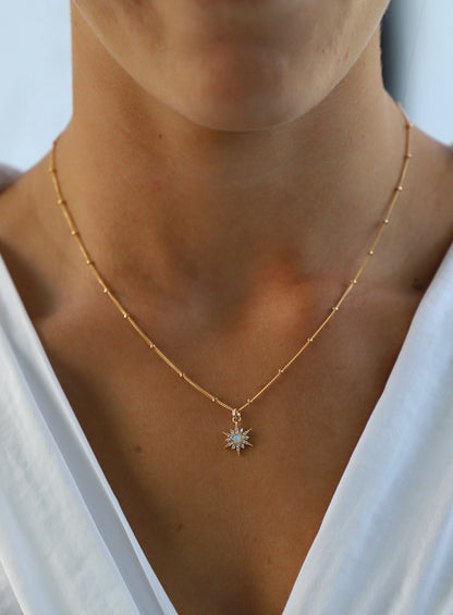 A gold chain necklace with a starburst pendant featuring blue stones hangs around the neck of a person, who is wearing a white top in a softly lit environment.