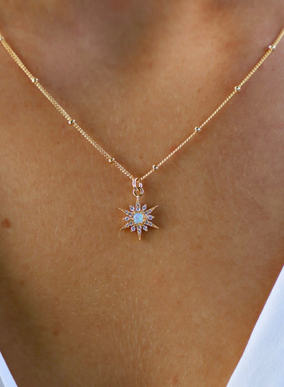 A gold necklace features a star-shaped pendant with a central opal and surrounding gemstones. It adorns a neck with a soft, tanned skin tone, contrasting with a white background.