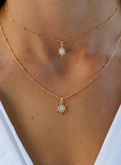 Two golden necklaces are layered around a neck, each featuring a star-shaped pendant with glimmering details. The skin underneath is smooth, and the context suggests a casual, elegant setting, highlighted by the soft lighting.