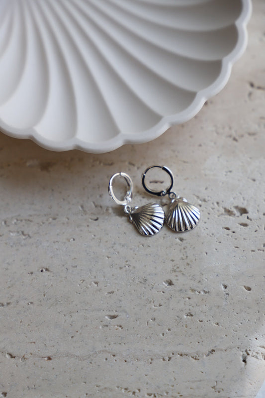 Silver shell-shaped earrings rest on a light-colored stone surface, accompanied by a white, ridged dish in the background, enhancing a minimalist and elegant presentation.