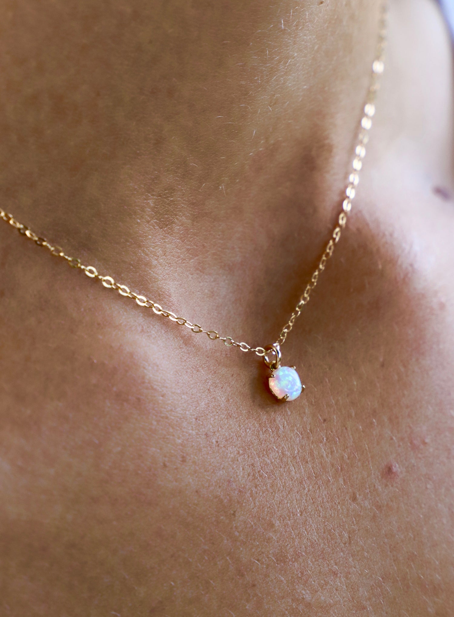 A delicate gold necklace rests against skin, featuring a small, iridescent opal pendant that reflects light, enhancing its subtle elegance in a soft, natural setting.