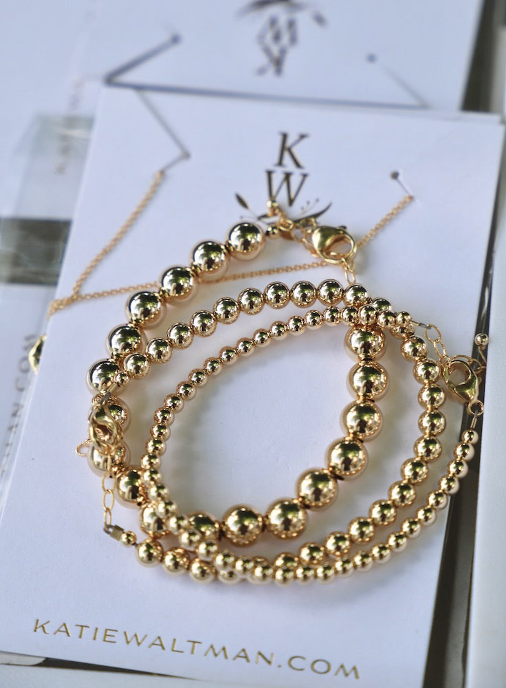 Three gold-tone bracelets, featuring various-sized spherical beads, are arranged on a branded card. The background includes faint text, and the overall setting suggests a display for jewelry.