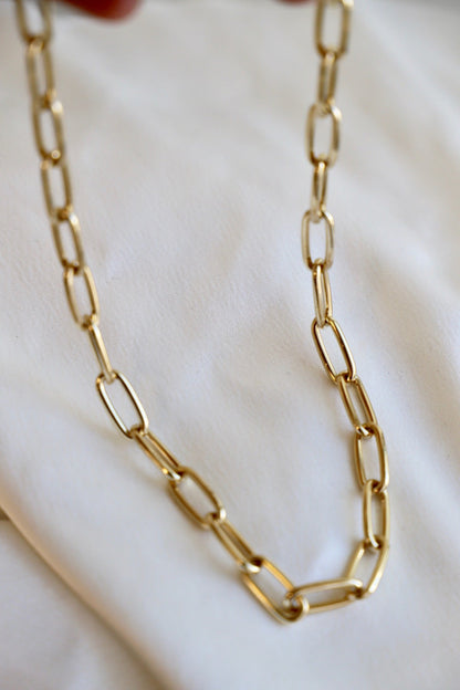 A gold chain necklace with elongated links is held above a soft, white fabric background, showcasing its shine and intricate design. The setting enhances the necklace's elegance.