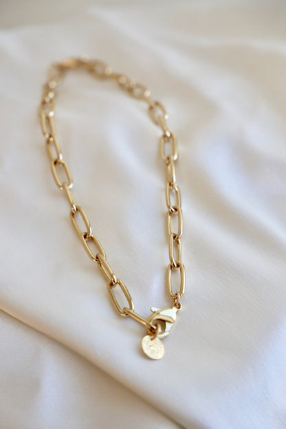 A golden chain necklace rests on a smooth, light-colored fabric. The necklace features elongated links and a decorative clasp, adding a stylish element to the soft background.