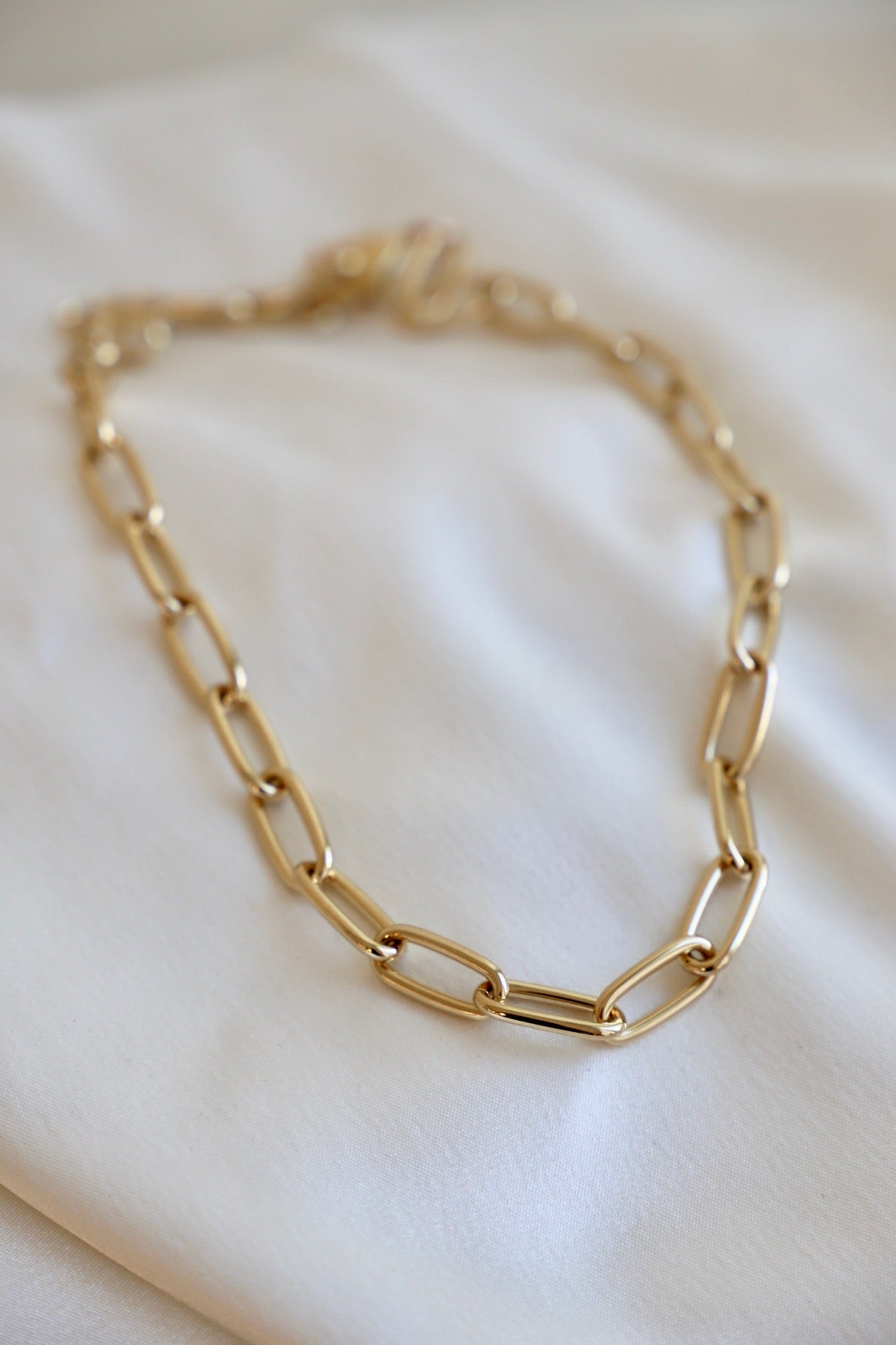 A gold chain necklace lays flat on a smooth, white fabric surface. The links are elongated and interlinked, capturing light softly in a minimalistic setting.