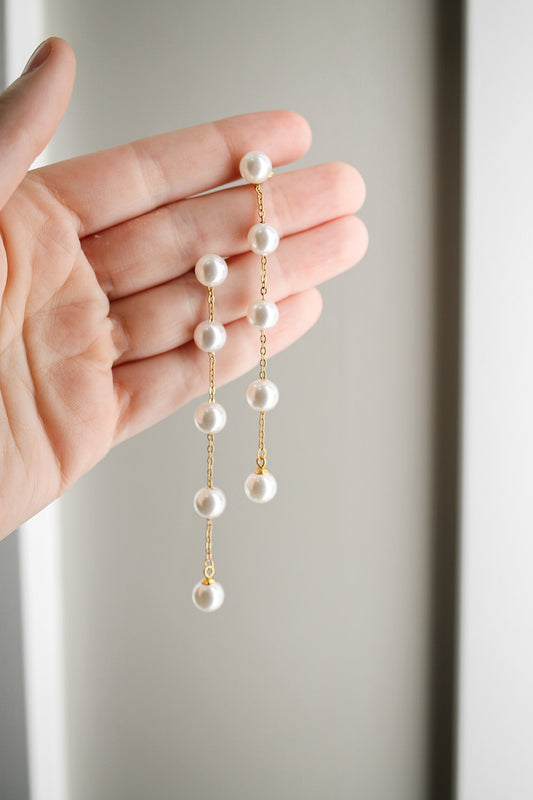 A hand holds a delicate gold chain earring adorned with multiple pearl beads, showcasing an elegant and refined style against a soft, neutral background.