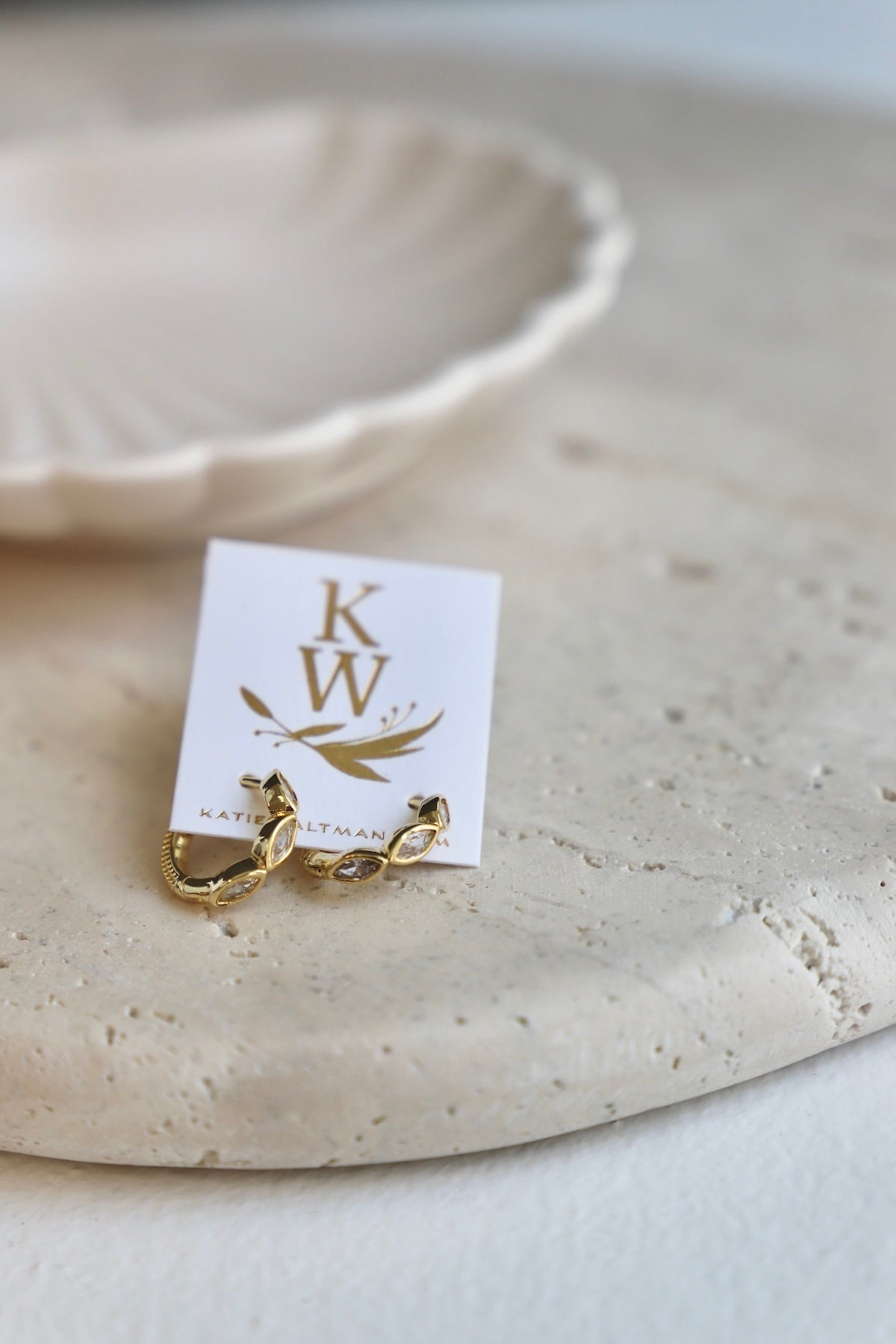 Two gold earrings are displayed on a textured, light-colored stone surface beside a white business card. A shallow, decorative dish is softly blurred in the background. The card reads "KW" and "KATIE ALT" beneath decorative elements.