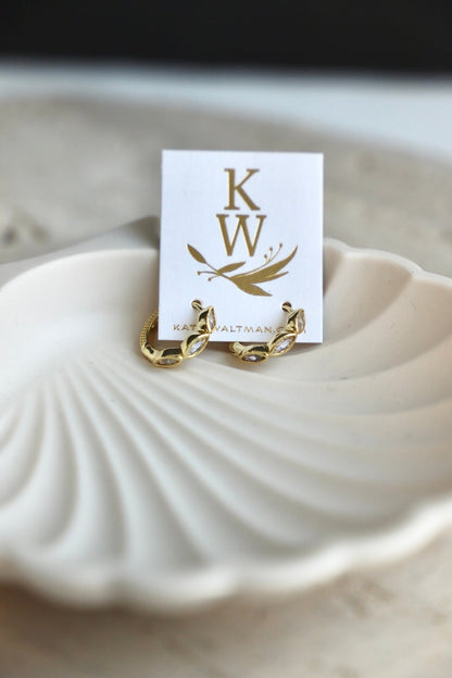 Gold jewelry earrings rest on a white, shell-shaped dish. A card behind them displays the initials "KW" and the name "KATE WALTMA." The background features soft, neutral tones.