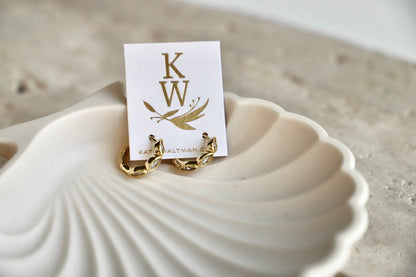 Gold earrings rest on a delicate shell-shaped dish, accompanied by a business card displaying the initials "KW" and floral designs, set against a softly blurred neutral background.