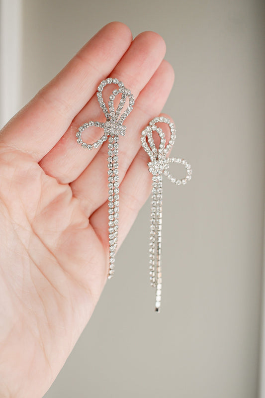 Two sparkling silver hairpins shaped like bows are held in a hand, showcasing their intricate design against a soft, neutral background. The pins feature a dangling chain of rhinestones.