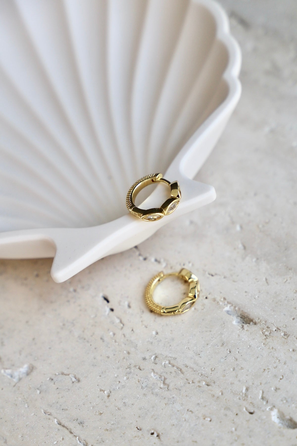 A white shell-shaped dish holds two gold earrings. One earring rests on the textured stone surface nearby. The scene is minimalistic and softly lit, emphasizing the jewelry's shine.