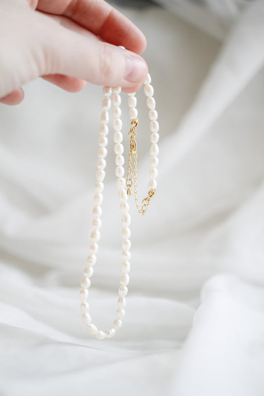 A hand holds a delicate pearl necklace, its shining beads elegantly draped while a golden clasp dangles. The background is softly blurred white fabric, enhancing the focus on the jewelry.