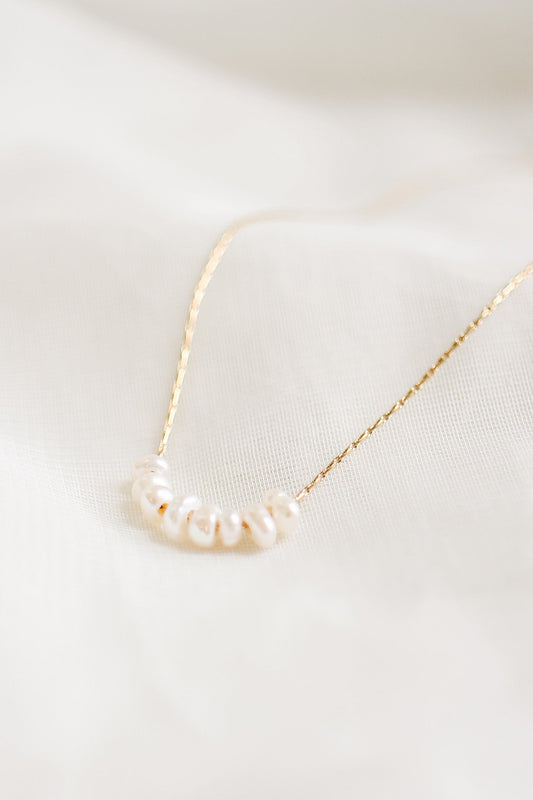 A delicate gold necklace with small, irregularly shaped pearls is elegantly displayed on a soft, light-colored fabric background, emphasizing its simplicity and charm.