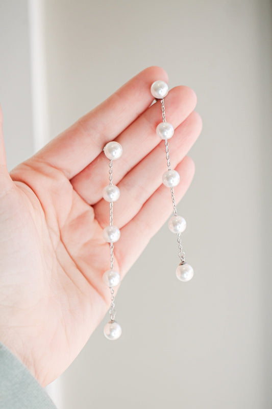 A hand holds a silver chain adorned with evenly spaced pearls, suspended vertically. The background features soft, neutral colors, suggesting a calm and elegant atmosphere.