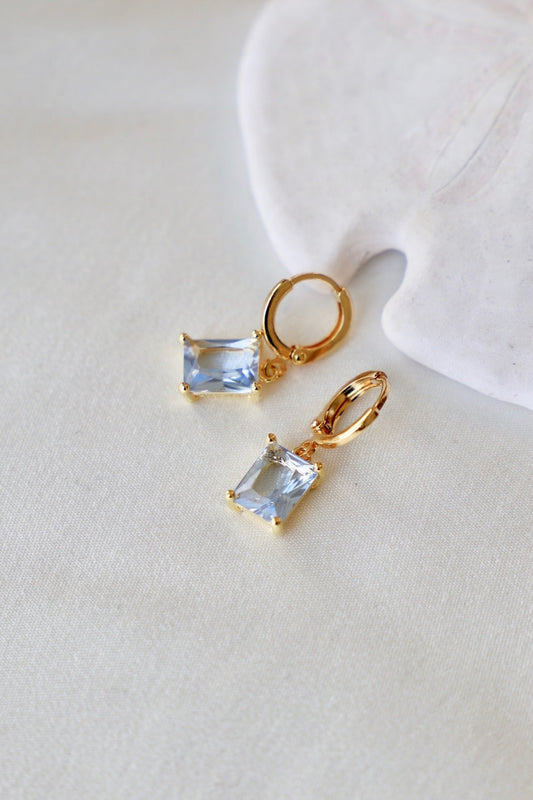 Two gold hoop earrings showcase rectangular blue gemstones, resting on a soft, light-colored fabric background that complements the jewelry's elegant design. The setting enhances their sparkle and allure.