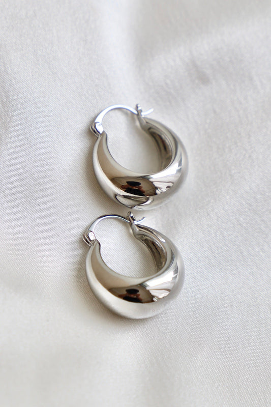 A pair of glossy silver hoop earrings rests on a smooth, light-colored fabric surface, reflecting light and displaying a curved, modern design with a simple clasp.