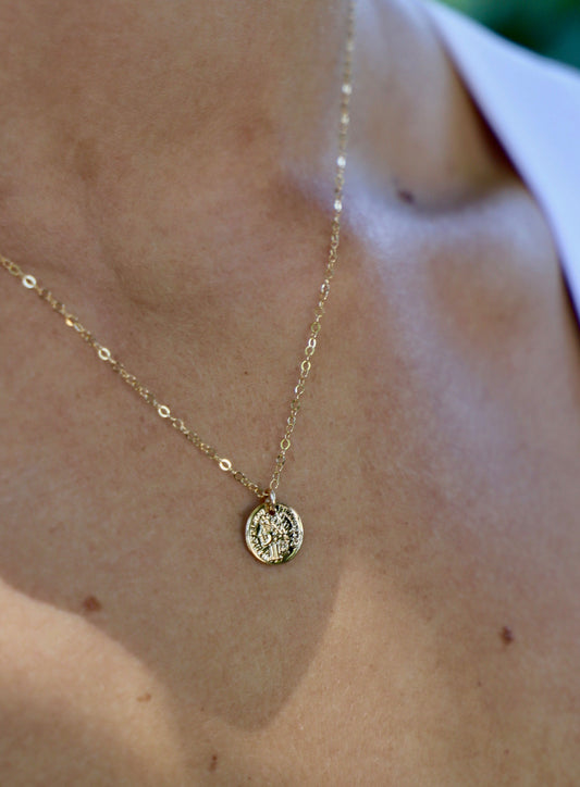 A gold necklace featuring a detailed circular pendant rests on skin, reflecting light. The background is softly blurred, suggesting an outdoor setting.