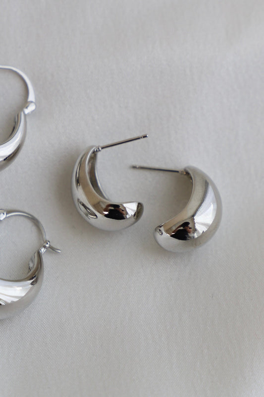 Silver earrings, shaped like crescent moons, are placed on a soft, light-colored fabric. Their shiny surfaces reflect light, highlighting their smooth curves and polished finish.