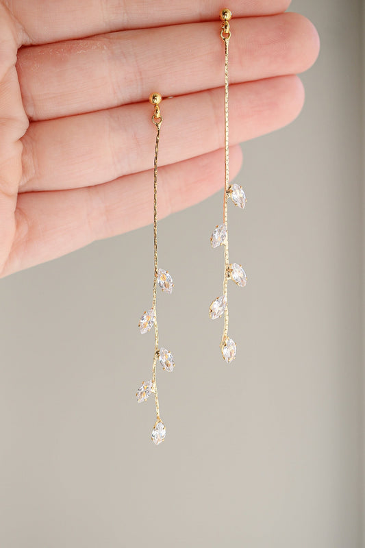 Dangling earrings with a gold chain, featuring clear crystal accents resembling leaves, are being held up by a hand against a soft, neutral background.