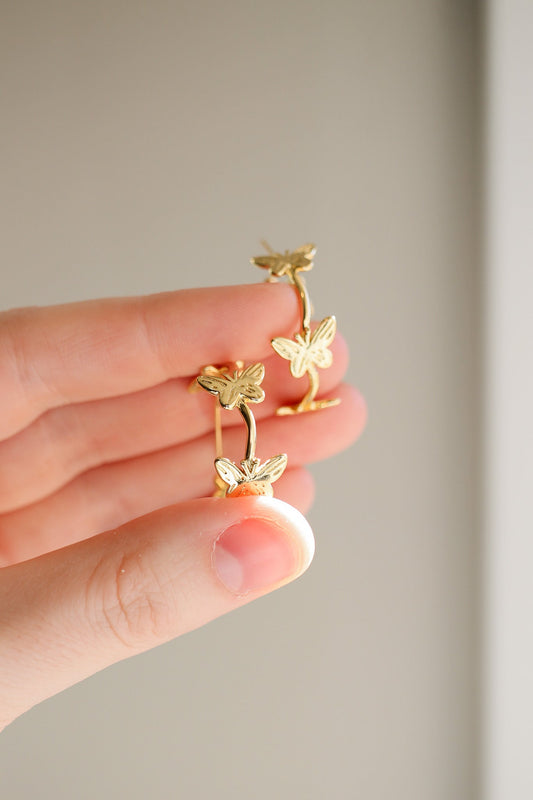 A hand holds a pair of gold butterfly earrings, displaying their delicate design against a neutral background, highlighting the intricate details of the butterflies and the shiny metal.