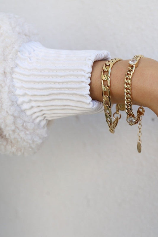 A hand features several gold bracelets layered over a white, ribbed sleeve. The background is a plain, light-colored wall, emphasizing the arm's accessories.