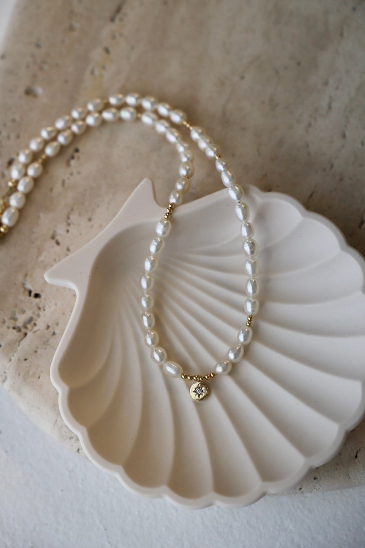 A delicate pearl necklace rests on a scalloped, white dish. The pearls are interspersed with small golden beads, and a gold pendant featuring a star design is centered.