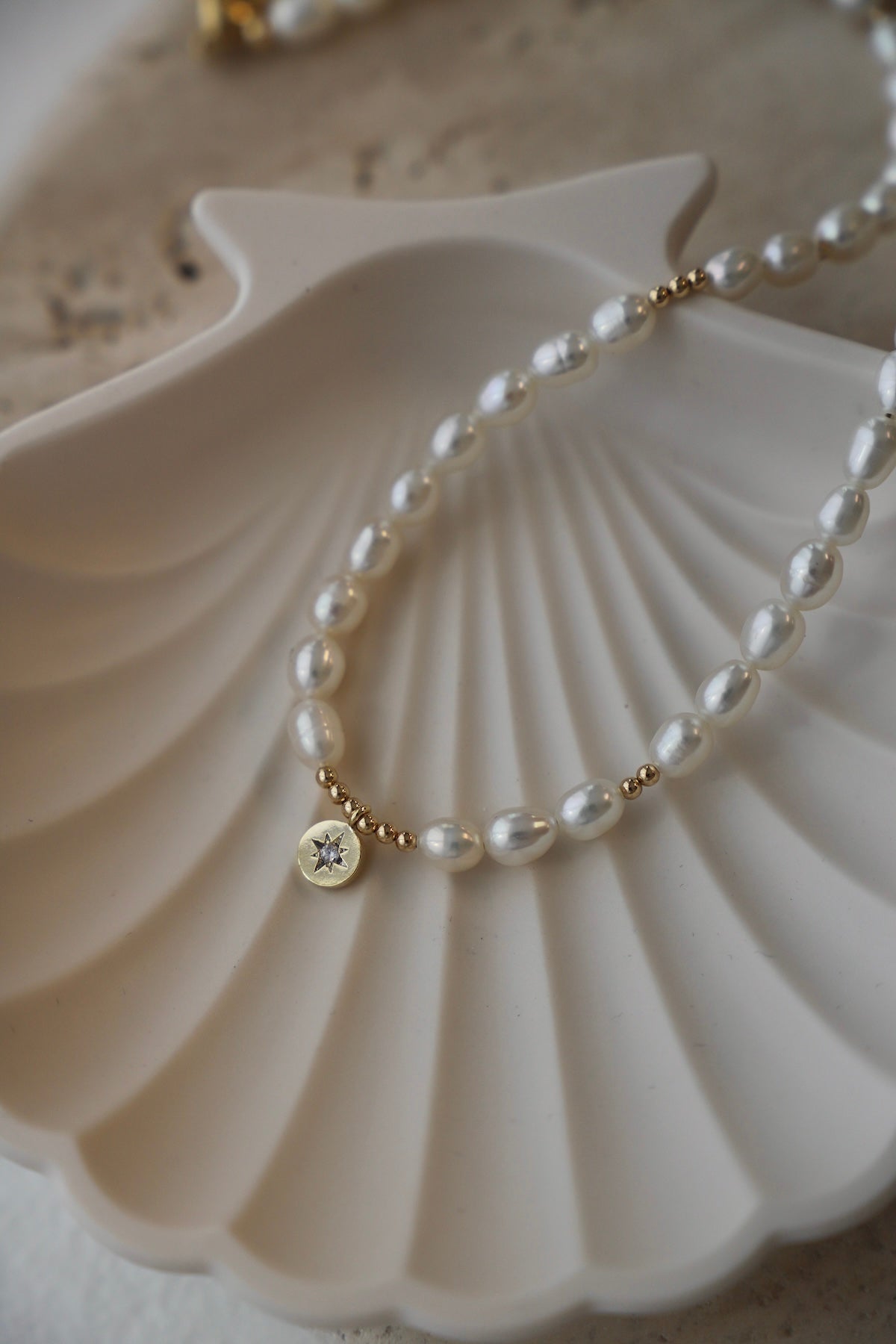 A delicate pearl necklace rests on a textured white shell dish. The necklace features pearl beads, interspersed with small gold accents, and includes a golden pendant with a star design.