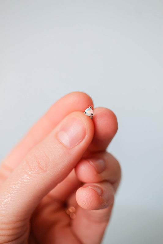 A hand holds a small silver stud earring with an iridescent gem, positioned against a soft, pale background, suggesting an atmosphere of delicacy and attention to detail.