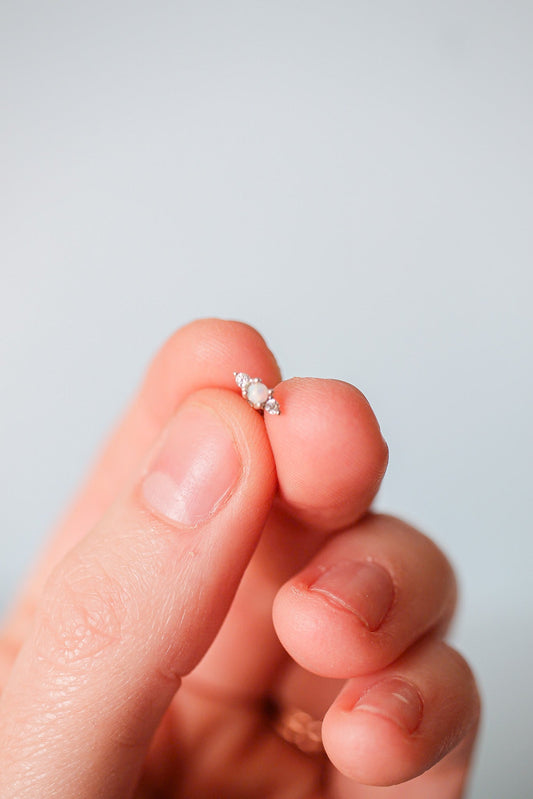 A hand holds a small, delicate earring featuring a central opal gemstone flanked by tiny diamonds, displayed against a light blue background.