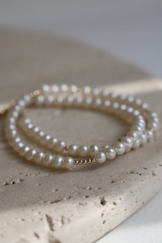 A delicate pearl bracelet coils gracefully on a textured stone surface, reflecting soft light. The background features muted tones, enhancing the elegance of the jewelry.