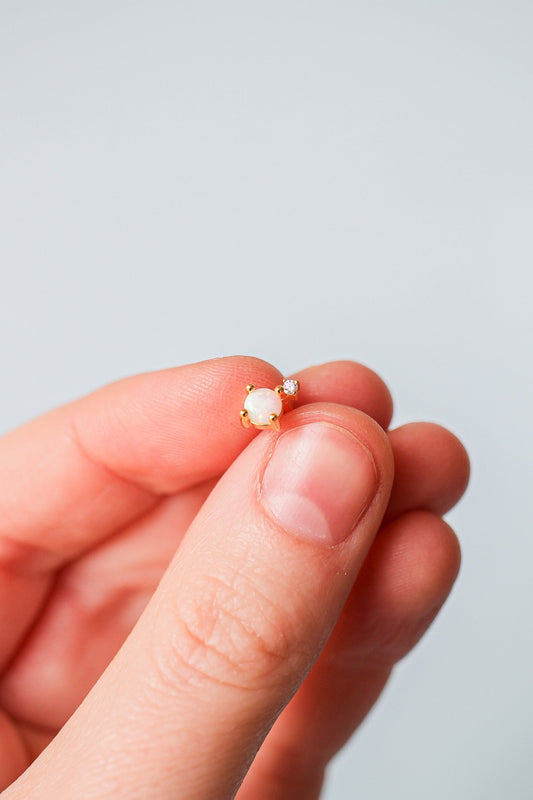 A hand holds a delicate gold earring featuring an opal stone surrounded by small gems, displayed against a soft, light gray background.