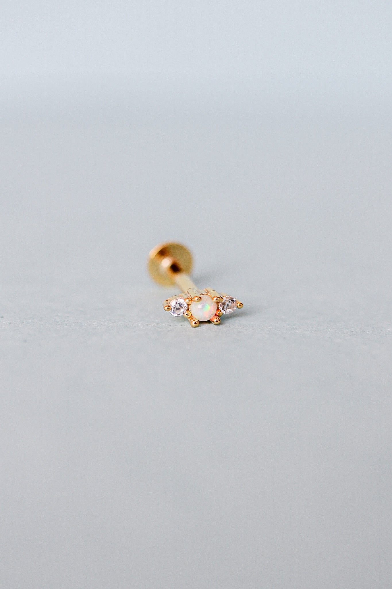 A gold stud earring rests on a light gray surface, featuring an opal stone flanked by two small diamonds, reflecting a soft shine in a minimalist, neutral setting.