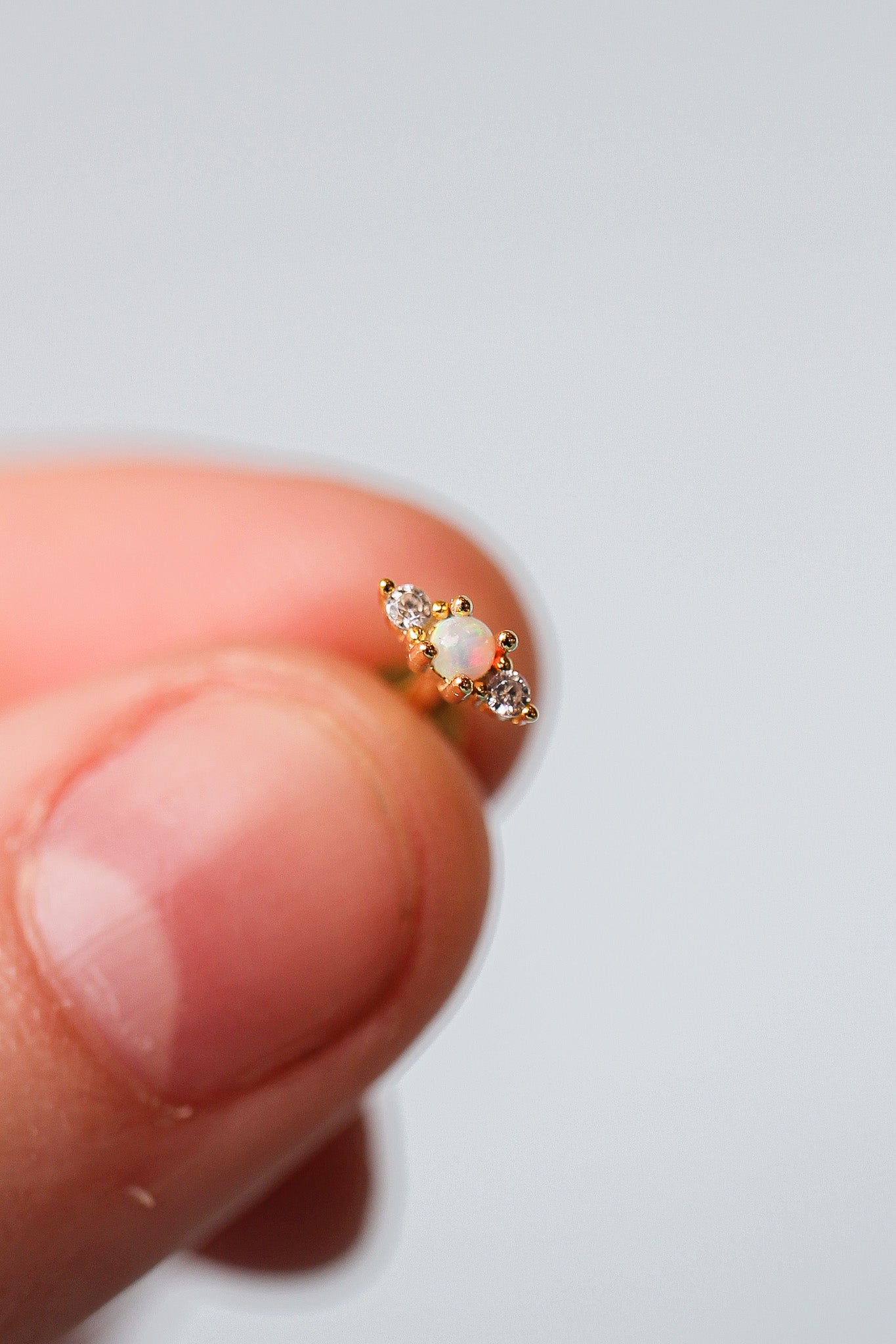 A hand gently holds a small, ornate earring featuring a central opal surrounded by two sparkling diamonds, against a soft, neutral background that enhances the earring's shine.