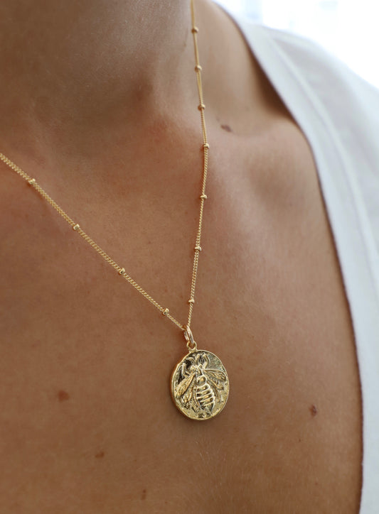 A golden bee pendant hangs from a delicate chain, resting on a skin tone. The background is softly blurred, suggesting an indoor, natural light setting, enhancing the pendant's shine.