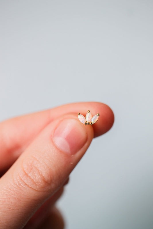 A hand holds a small, delicate piece of jewelry featuring three opal stones arranged like petals, against a soft, neutral background that emphasizes the object.