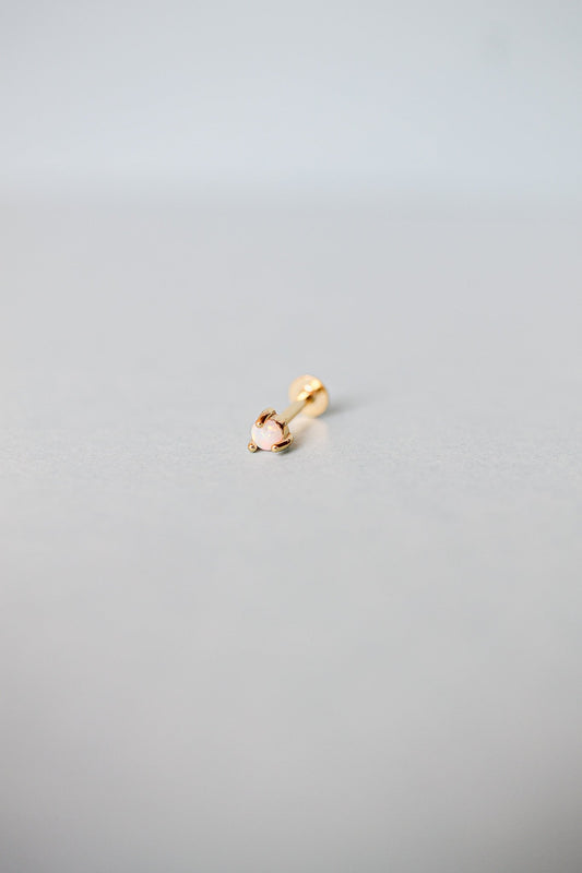 A small gold earring lies on a light gray surface, featuring a round opal stone at the end. The setting has a delicate design, suggesting elegance in its simplicity.