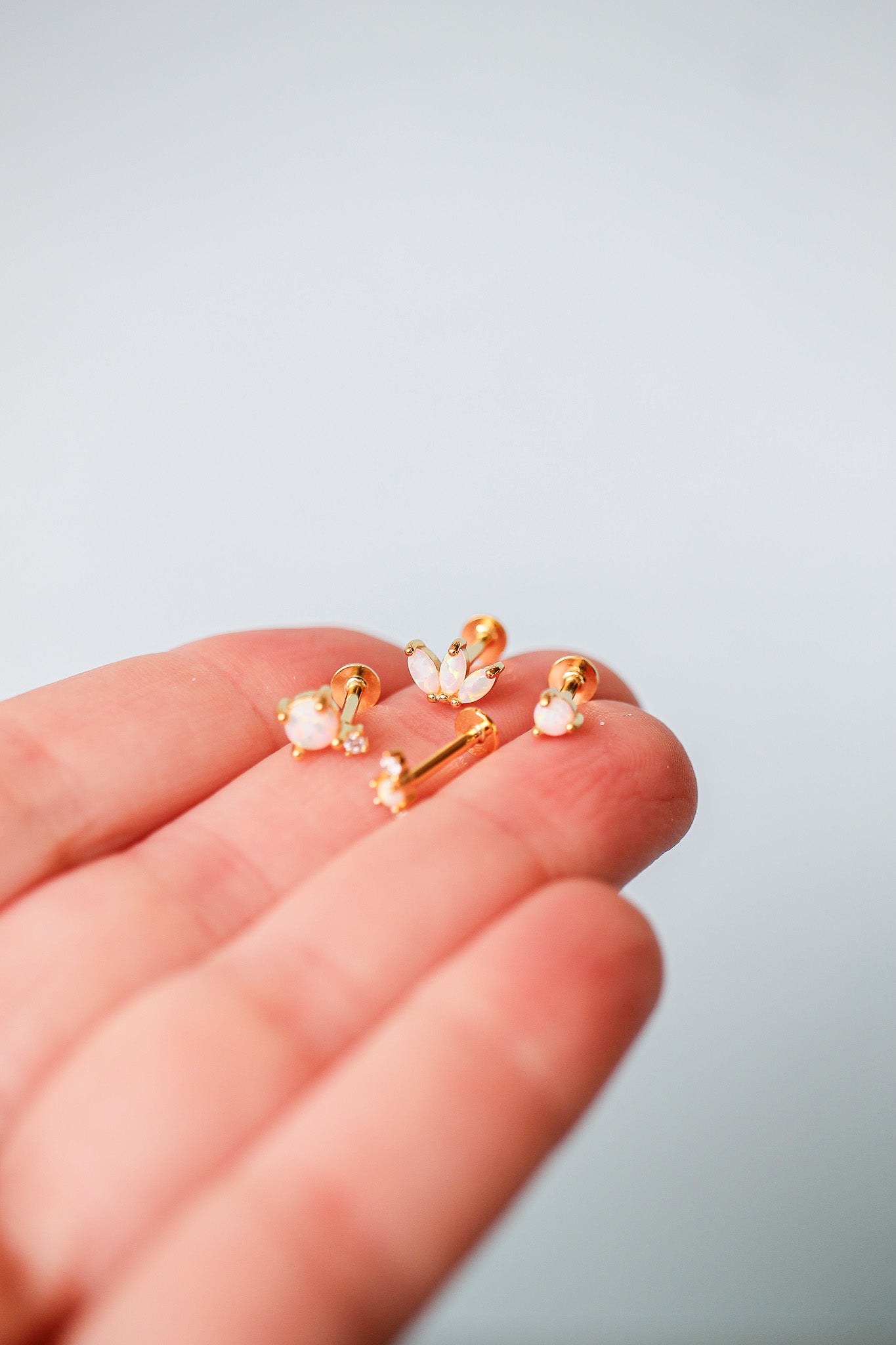 A hand holds several pieces of jewelry, including gold earrings with opal accents and a decorative gold bar. The background is a neutral, soft color, emphasizing the jewelry.