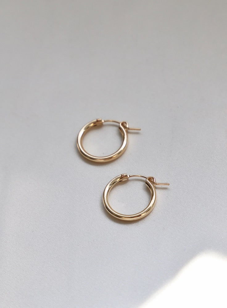 Two gold hoop earrings are resting on a soft, light-colored fabric, showcasing their shiny surface and circular design. The ambient light highlights their smooth edges.