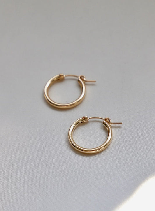 Two gold hoop earrings rest on a soft, light-colored fabric. The earrings are circular with a smooth surface and a small clasp, appearing elegant and simple in design.