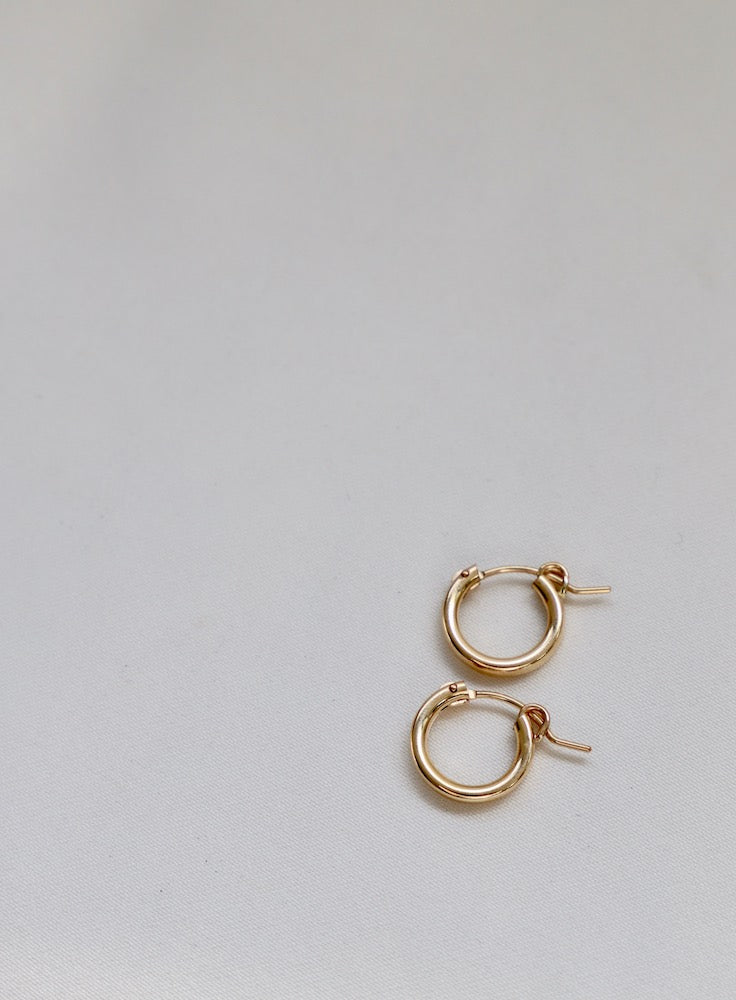 Two gold hoop earrings are placed on a light, neutral background. The larger earring hangs above a smaller one, showcasing their polished surfaces and simple design.