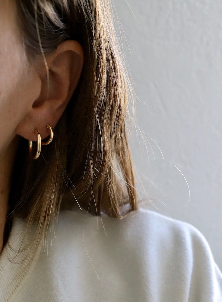 Gold hoop earrings adorn the ear of a person with long, light brown hair. The background is a smooth, light-colored wall. The person wears a white sweatshirt.