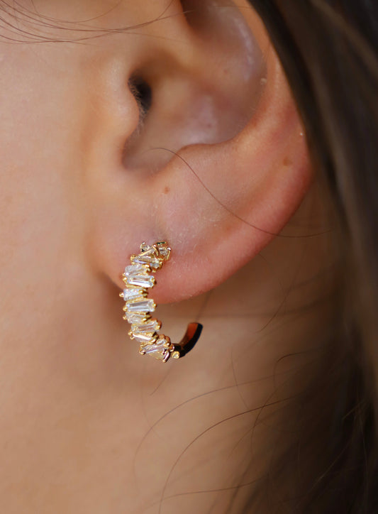 Gold hoop earrings adorned with several sparkling, elongated gemstones are clipped onto the outer part of a person's ear, surrounded by light skin and dark hair.