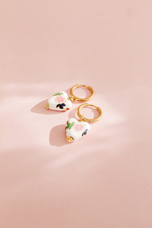 Two heart-shaped earrings with floral designs hang from gold hoop clasps, resting on a soft, pink surface, illuminated by gentle light that creates delicate shadows.