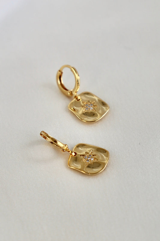 Gold earrings with a textured, square-shaped pendant featuring a star design adorned with small stones are displayed against a light, neutral fabric background.