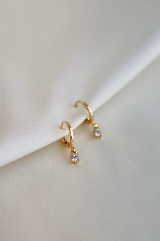 Gold hoop earrings with small diamond-like stones are resting on a soft, cream-colored fabric. Their elegant design catches light, emphasizing their shine against the textured background.