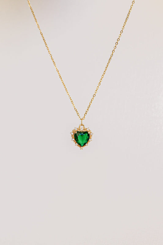 A gold chain necklace features a heart-shaped green gemstone pendant surrounded by small sparkling accents, suspended against a soft, light-colored background.